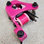 A pink coil tattoo machine resting on a concrete surface.
