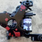 Close-up of a black tattoo machine with red splatter design and a grip featuring a crow image.